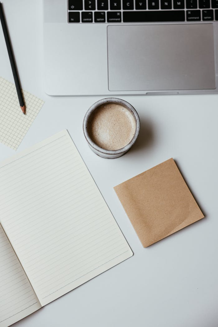 Top view of stylish workspace featuring a laptop, coffee, notepad, and pencil on a white surface.