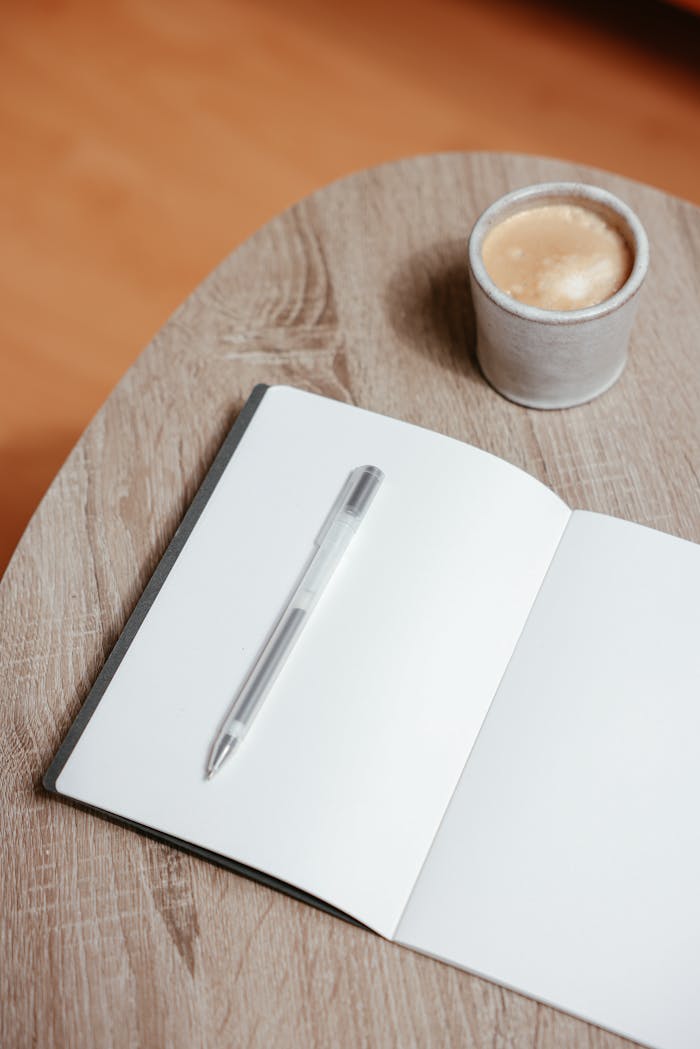 An open notebook with a pen beside a cup of coffee on a wooden table.